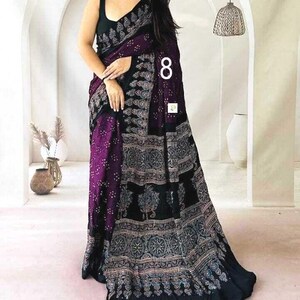May include: A black and deep purple saree featuring a sleeveless blouse and a long, flowing drape. The fabric is adorned with intricate, repeating white and light-colored floral and geometric patterns throughout the body and the wide border.