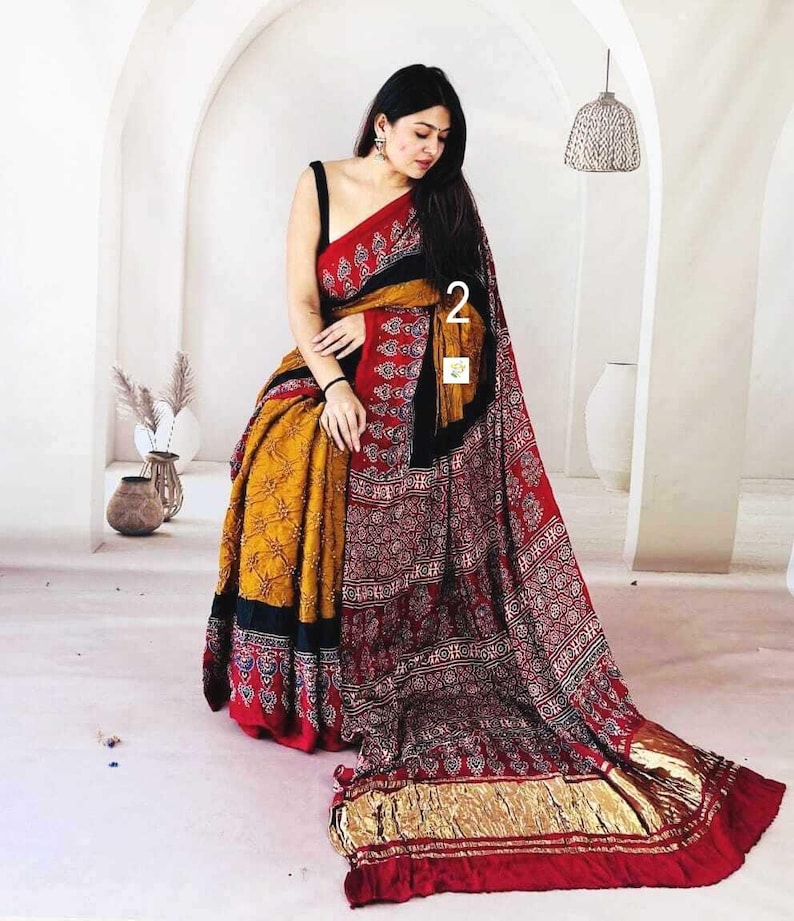 May include: A traditional saree featuring a vibrant mustard yellow section with a dark floral pattern, contrasted by a deep red section adorned with intricate white and black block-printed motifs, finished with a metallic gold border and black trim.