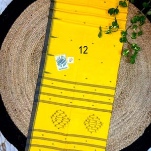 May include: A bright yellow textile featuring a dark gray border with thin horizontal stripes. The fabric displays small scattered dots, two large diamond-shaped patterns, and a white label with the text 12 and Handloom. It has a fringed edge.