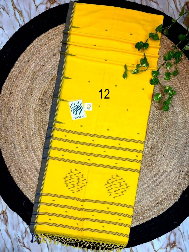 May include: A bright yellow textile featuring a dark gray border with thin horizontal stripes. The fabric displays small scattered dots, two large diamond-shaped patterns, and a white label with the text 12 and Handloom. It has a fringed edge.