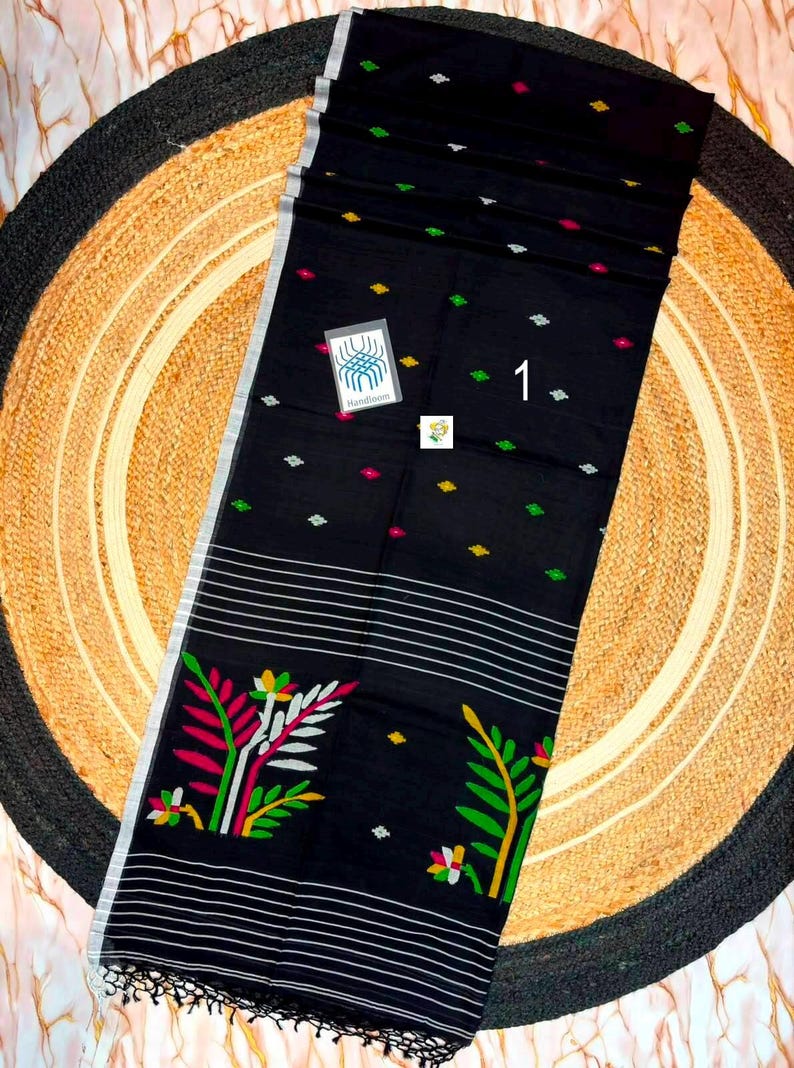 May include: A black textile featuring a pattern of small green, yellow, and pink diamond shapes, horizontal white stripes, and large floral motifs in pink, green, and white. A small tag displays the number 1 and a logo with the text Handloom.
