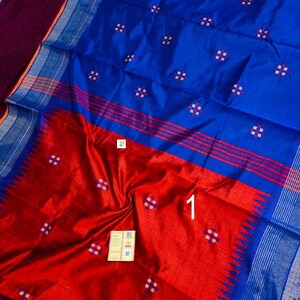 May include: A silk saree in blue and red, featuring a detailed pattern. The saree has a colour block design with a blue upper section and a red lower section. It includes small, square, white and red floral motifs.