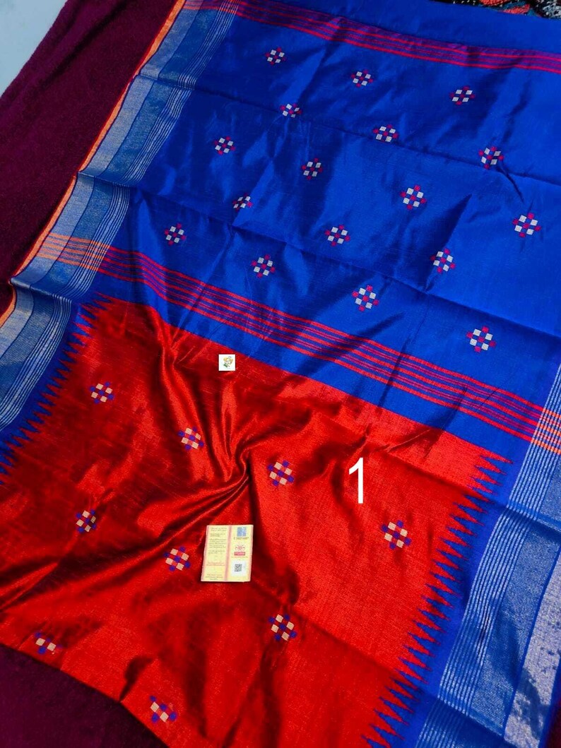 May include: A silk saree in blue and red, featuring a detailed pattern. The saree has a colour block design with a blue upper section and a red lower section. It includes small, square, white and red floral motifs.
