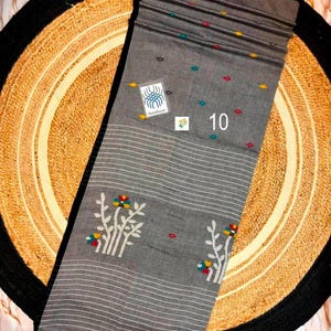 May include: A grey fabric textile featuring white horizontal stripes, small scattered multi-colored floral motifs, and larger white floral branch patterns. A square label reads Handloom, and the number 10 is printed in white on the fabric.