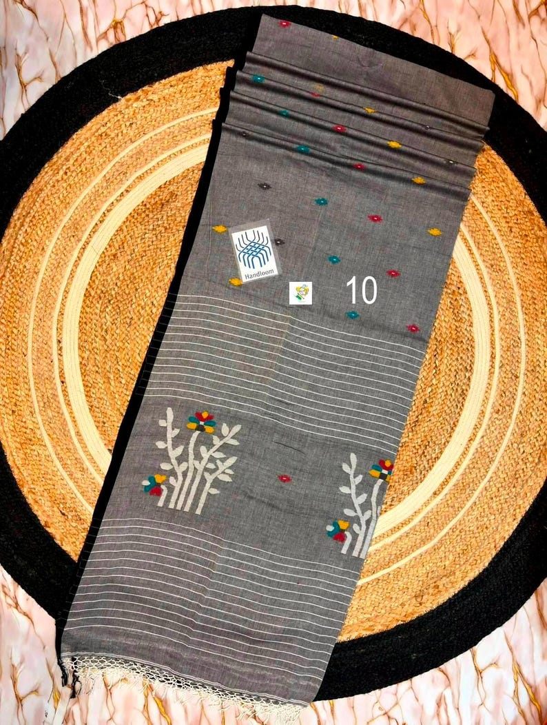 May include: A grey fabric textile featuring white horizontal stripes, small scattered multi-colored floral motifs, and larger white floral branch patterns. A square label reads Handloom, and the number 10 is printed in white on the fabric.