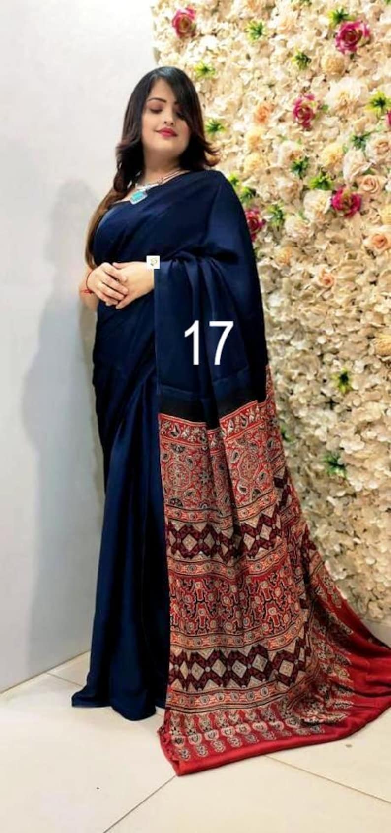 Puede incluir: Un sari azul marino oscuro con un cuerpo liso y de color s&oacute;lido, y un pallu ancho y contrastante con intrincados patrones geom&eacute;tricos y florales en rojo, negro y blanco. El n&uacute;mero 17 est&aacute; impreso en blanco en el centro de la tela.
