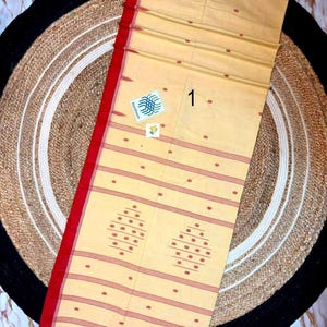May include: A light beige cotton textile featuring a bold red border, horizontal red stripes, and small red dot patterns. The fabric includes two larger diamond-shaped motifs made of red dots and a small white square tag with the number 1.