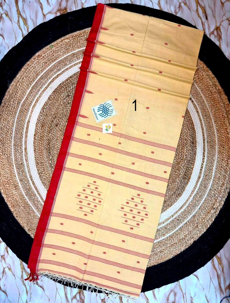 May include: A light beige cotton textile featuring a bold red border, horizontal red stripes, and small red dot patterns. The fabric includes two larger diamond-shaped motifs made of red dots and a small white square tag with the number 1.