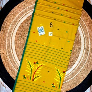 May include: A mustard yellow textile featuring a dark green border and thin horizontal green stripes. The fabric is decorated with small red, green, and yellow diamond-shaped motifs and larger yellow and green leaf patterns. A tag labeled 8 is visible.