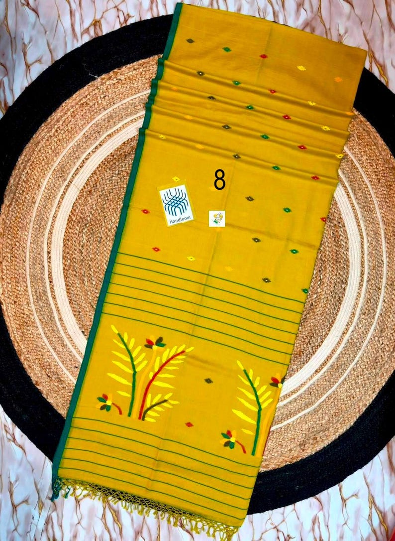 May include: A mustard yellow textile featuring a dark green border and thin horizontal green stripes. The fabric is decorated with small red, green, and yellow diamond-shaped motifs and larger yellow and green leaf patterns. A tag labeled 8 is visible.