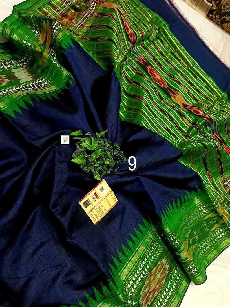 May include: A dark navy blue fabric with a vibrant green border featuring intricate ikat patterns and a serrated edge. The textile displays a woven texture, a small green plant sprig, the number 9, and a rectangular product tag with text.