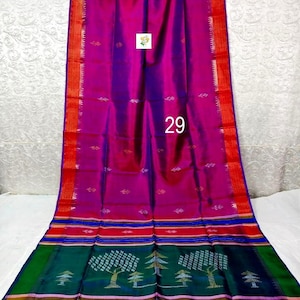 Puede incluir: Un sari magenta y morado con un borde rojo con detalles dorados y peque&ntilde;os motivos de hojas. La secci&oacute;n inferior muestra un panel verde oscuro con patrones de &aacute;rboles tejidos, rayas horizontales en azul, rojo y dorado, y el n&uacute;mero 29 en blanco.