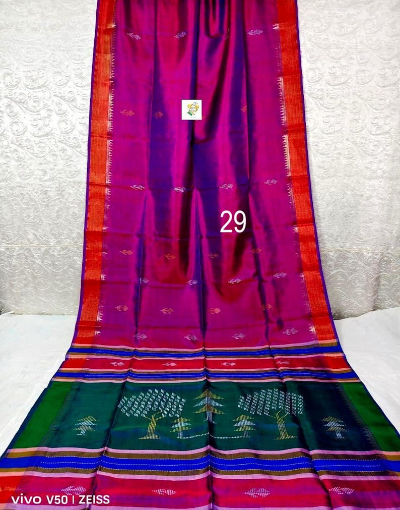 Puede incluir: Un sari magenta y morado con un borde rojo con detalles dorados y peque&ntilde;os motivos de hojas. La secci&oacute;n inferior muestra un panel verde oscuro con patrones de &aacute;rboles tejidos, rayas horizontales en azul, rojo y dorado, y el n&uacute;mero 29 en blanco.