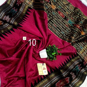 May include: A magenta saree featuring a black border with a gold metallic stripe and a triangular pattern. The fabric displays a black section with multicolored ikat motifs and gold stripes. A small white tag with the number 10 is attached.