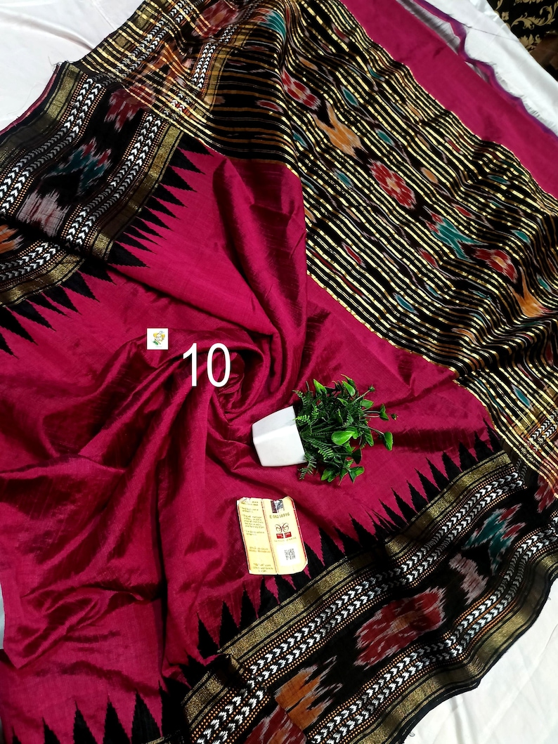 May include: A magenta saree featuring a black border with a gold metallic stripe and a triangular pattern. The fabric displays a black section with multicolored ikat motifs and gold stripes. A small white tag with the number 10 is attached.