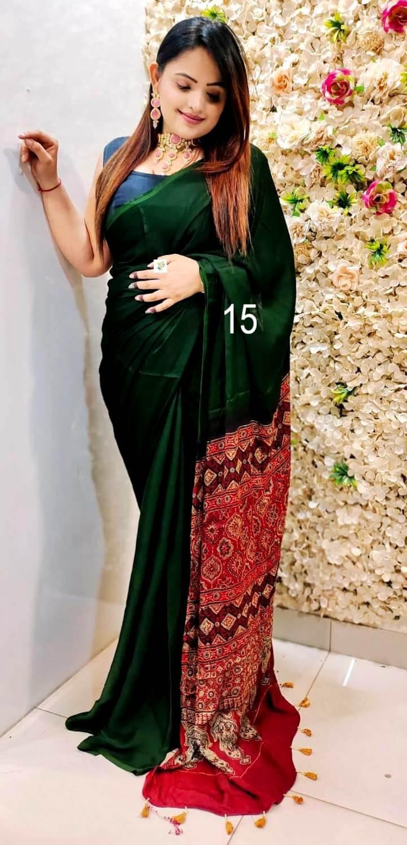 Puede incluir: Un sari de color verde oscuro con un drapeado liso y un extremo rojo en contraste que presenta intrincados patrones geom&eacute;tricos en blanco y dorado. La prenda incluye una blusa azul oscuro sin mangas, un collar tipo gargantilla y pendientes a juego.