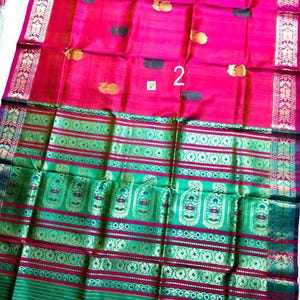 May include: A vibrant fuchsia pink saree featuring scattered gold and dark green floral motifs. The fabric includes a wide, ornate border in emerald green and gold with intricate paisley patterns and horizontal stripes. The number 2 is visible.