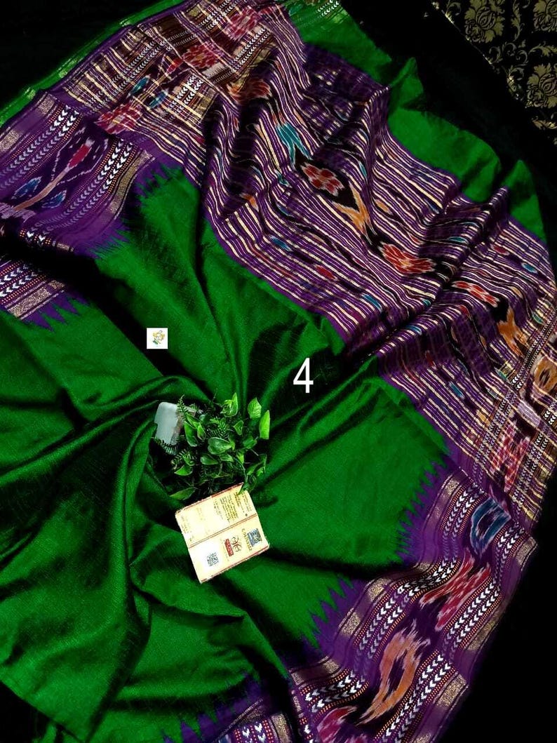 May include: A vibrant emerald green saree featuring a wide purple border with intricate ikat patterns in white, red, and yellow. The fabric displays a smooth texture with a prominent number 4 printed on the surface and a small attached product tag.