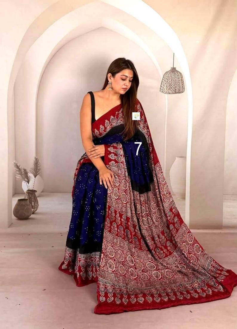 May include: A saree featuring a deep blue central panel with a white dotted pattern, bordered by a wide red section adorned with intricate white and black floral motifs. The garment includes a black sleeveless blouse and a prominent number 7.