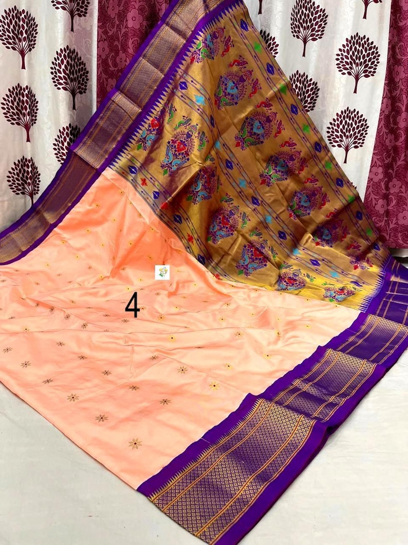 Op de afbeelding: Een perzikkleurige sari met een brede paarse rand met gouden geometrische patronen. De pallu is goudkleurig met kleurrijke pauwenmotieven. Het hoofdgedeelte heeft kleine verspreide bloemenborduursels en het cijfer 4 is op de stof gedrukt.