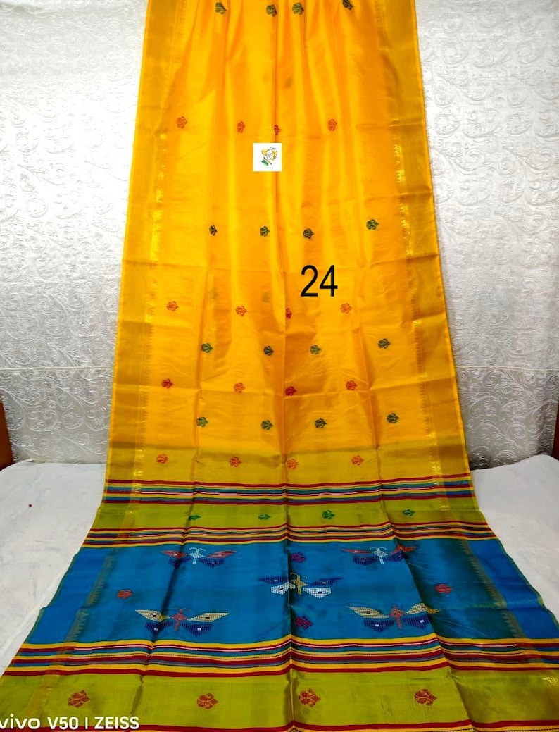 Puede incluir: Un sari de color amarillo brillante con peque&ntilde;os motivos florales rojos y verdes, con un borde ancho que contiene rayas horizontales en verde, rojo y azul. La secci&oacute;n azul muestra patrones de mariposas. El n&uacute;mero 24 est&aacute; impreso en la tela.