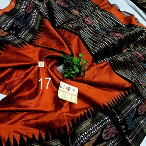 May include: A rust-orange saree featuring a wide black border with intricate white geometric patterns and a black pallu section with vertical stripes and floral motifs. The fabric has a subtle sheen, and the number 17 is printed in white.