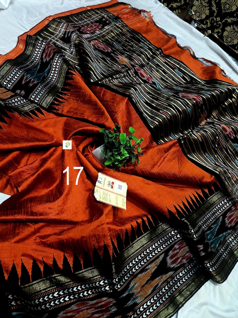May include: A rust-orange saree featuring a wide black border with intricate white geometric patterns and a black pallu section with vertical stripes and floral motifs. The fabric has a subtle sheen, and the number 17 is printed in white.