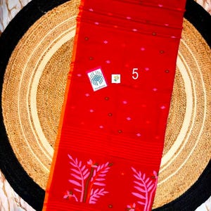 May include: A vibrant red textile featuring a repeating pattern of small pink and brown diamond-shaped motifs. The lower section displays a horizontal striped border with large pink leaf illustrations and a fringed edge. A small tag labeled 5 is visible.