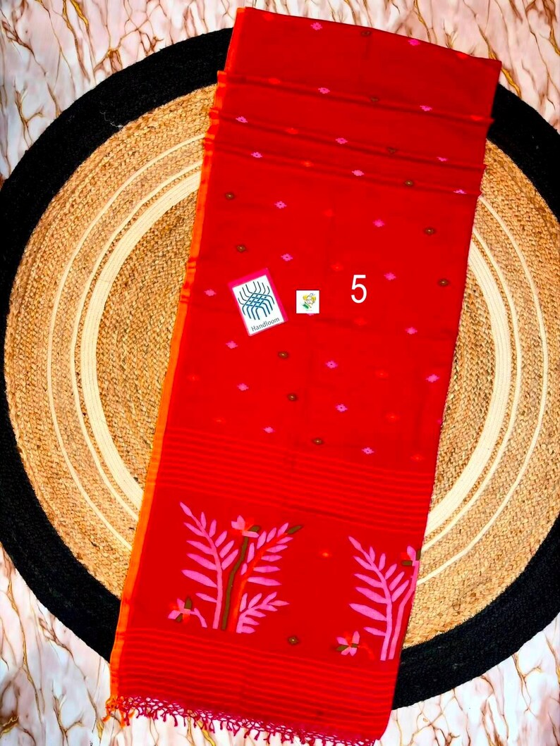 May include: A vibrant red textile featuring a repeating pattern of small pink and brown diamond-shaped motifs. The lower section displays a horizontal striped border with large pink leaf illustrations and a fringed edge. A small tag labeled 5 is visible.