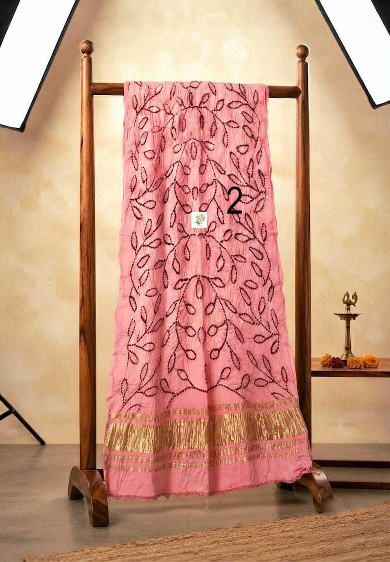 May include: A light pink fabric drape featuring a dark maroon embroidered leaf pattern and a wide metallic gold border at the bottom. The fabric has a crinkled texture and displays the number 2 in black print near the center of the material.