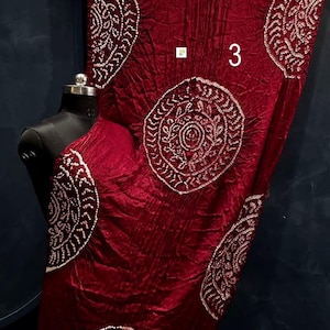 May include: A deep red fabric with a crinkled texture featuring large circular white patterns and the number 3 printed on the surface. The fabric is draped over a black mannequin torso and includes a metallic gold border at the bottom edge.