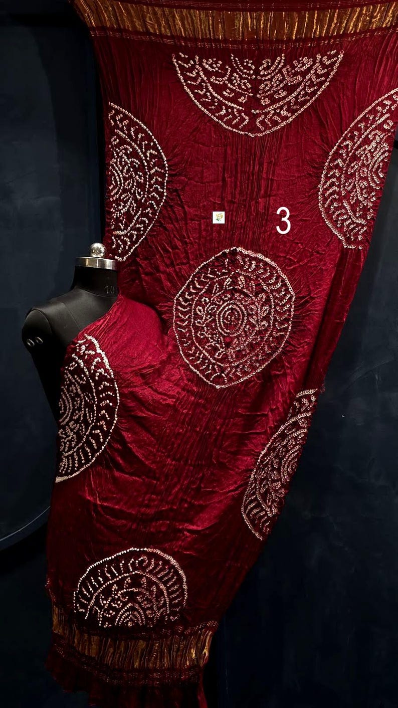 May include: A deep red fabric with a crinkled texture featuring large circular white patterns and the number 3 printed on the surface. The fabric is draped over a black mannequin torso and includes a metallic gold border at the bottom edge.