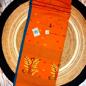 May include: An orange fabric textile featuring a blue border, small green and red diamond-shaped patterns, and a large floral motif at the bottom. The fabric includes the number 4, a small square tag, and a rectangular tag with the text Handloom.