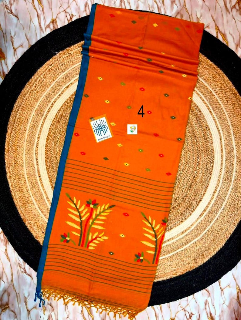 May include: An orange fabric textile featuring a blue border, small green and red diamond-shaped patterns, and a large floral motif at the bottom. The fabric includes the number 4, a small square tag, and a rectangular tag with the text Handloom.