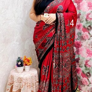 May include: A red saree featuring a black and white block-print pattern with floral motifs, a solid black border, and a metallic gold pleated hem. The garment is paired with a sleeveless black blouse and includes the number 4 printed on the fabric.
