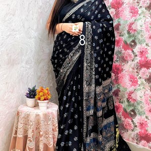 May include: A black saree featuring a repeating white floral print, a wide decorative border with intricate geometric and floral patterns in blue and white, and a metallic gold pleated end piece, worn with a matching black sleeveless blouse.