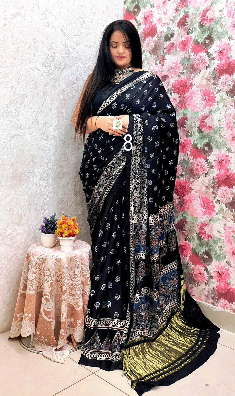 May include: A black saree featuring a repeating white floral print, a wide decorative border with intricate geometric and floral patterns in blue and white, and a metallic gold pleated end piece, worn with a matching black sleeveless blouse.