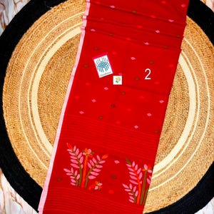 May include: A vibrant red textile featuring a light pink border and small scattered geometric motifs in pink and brown. The lower section displays two stylized floral patterns in pink, orange, and green. A small white tag displays the number 2.