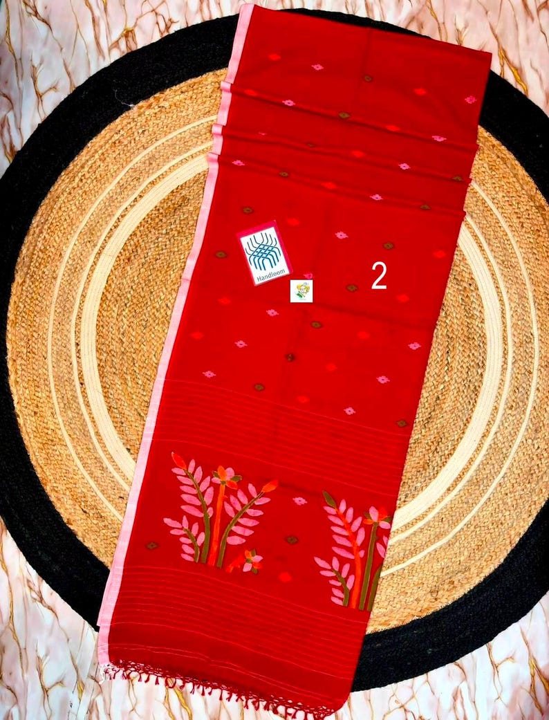 May include: A vibrant red textile featuring a light pink border and small scattered geometric motifs in pink and brown. The lower section displays two stylized floral patterns in pink, orange, and green. A small white tag displays the number 2.