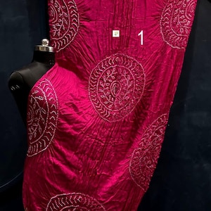 May include: A vibrant magenta fabric wrap featuring large circular white tie-dye patterns and intricate detailing. The textile has a crinkled texture, a metallic gold border at the top and bottom edges, and the number 1 printed in white on the surface.