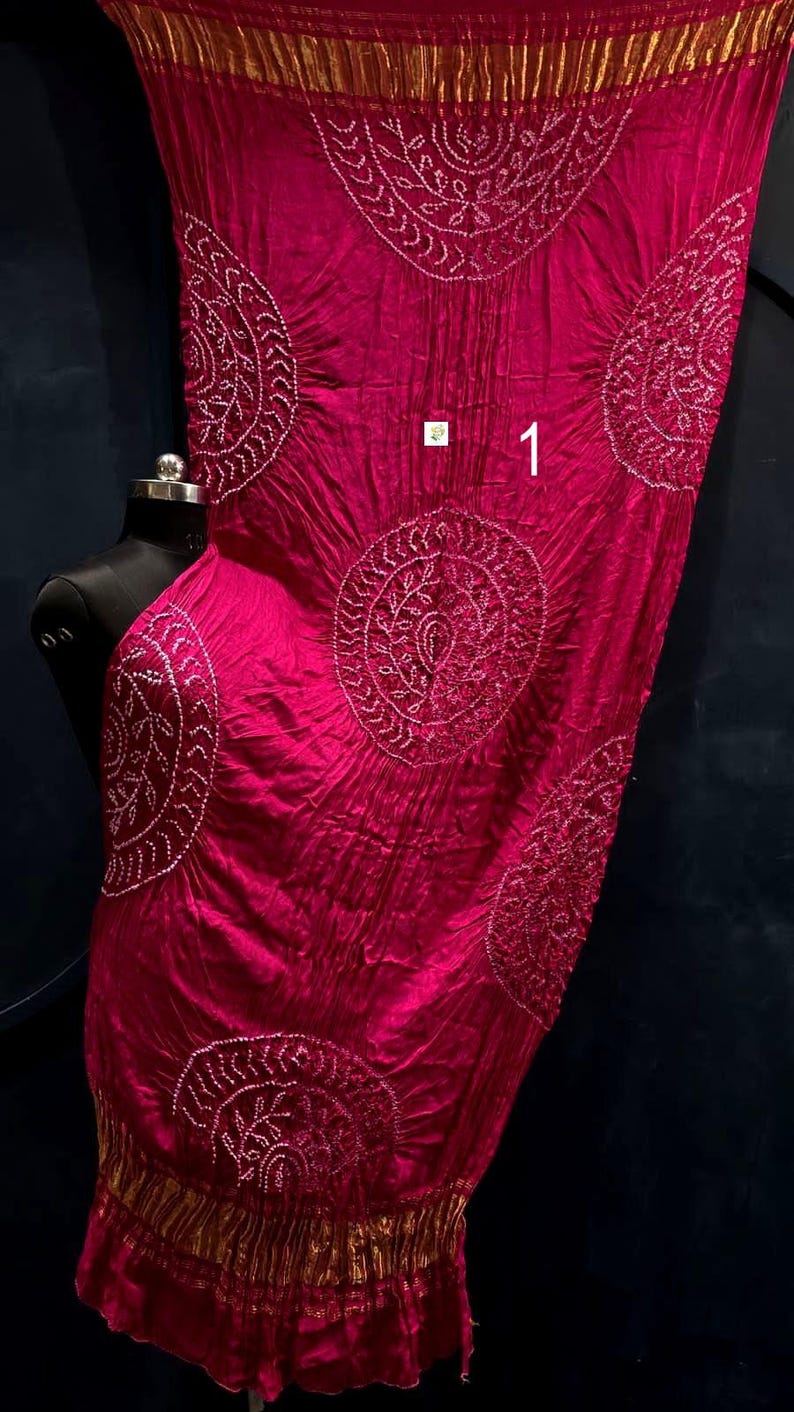May include: A vibrant magenta fabric wrap featuring large circular white tie-dye patterns and intricate detailing. The textile has a crinkled texture, a metallic gold border at the top and bottom edges, and the number 1 printed in white on the surface.