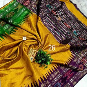 May include: A mustard yellow saree featuring a wide purple border with intricate ikat patterns and a sharp triangular edge design. A vibrant green section displays floral motifs, while the number 26 is printed on the fabric near a small tag.