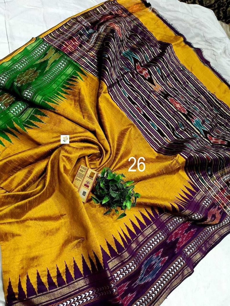 May include: A mustard yellow saree featuring a wide purple border with intricate ikat patterns and a sharp triangular edge design. A vibrant green section displays floral motifs, while the number 26 is printed on the fabric near a small tag.