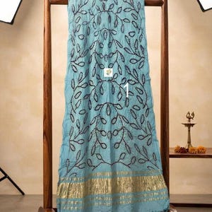 May include: A light blue fabric drape featuring a dark, intricate leaf-patterned embroidery throughout the center. The bottom edge is finished with a wide, metallic gold band with horizontal stripes and a frayed, textured hemline.