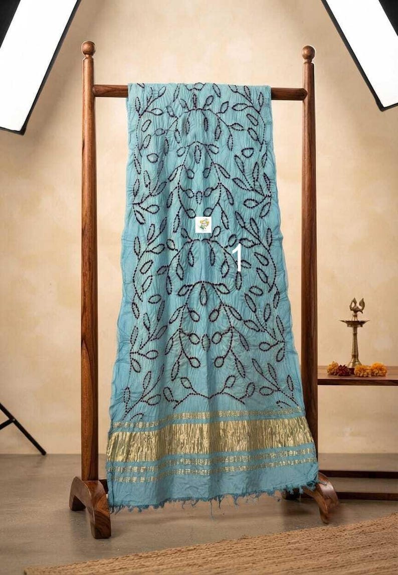 May include: A light blue fabric drape featuring a dark, intricate leaf-patterned embroidery throughout the center. The bottom edge is finished with a wide, metallic gold band with horizontal stripes and a frayed, textured hemline.