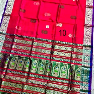 May include: A vibrant red saree featuring a wide green border with intricate gold and multicolored paisley patterns. The fabric displays small floral motifs across the main body and a prominent number 10 printed on the surface. The edges are finished with a purple trim.