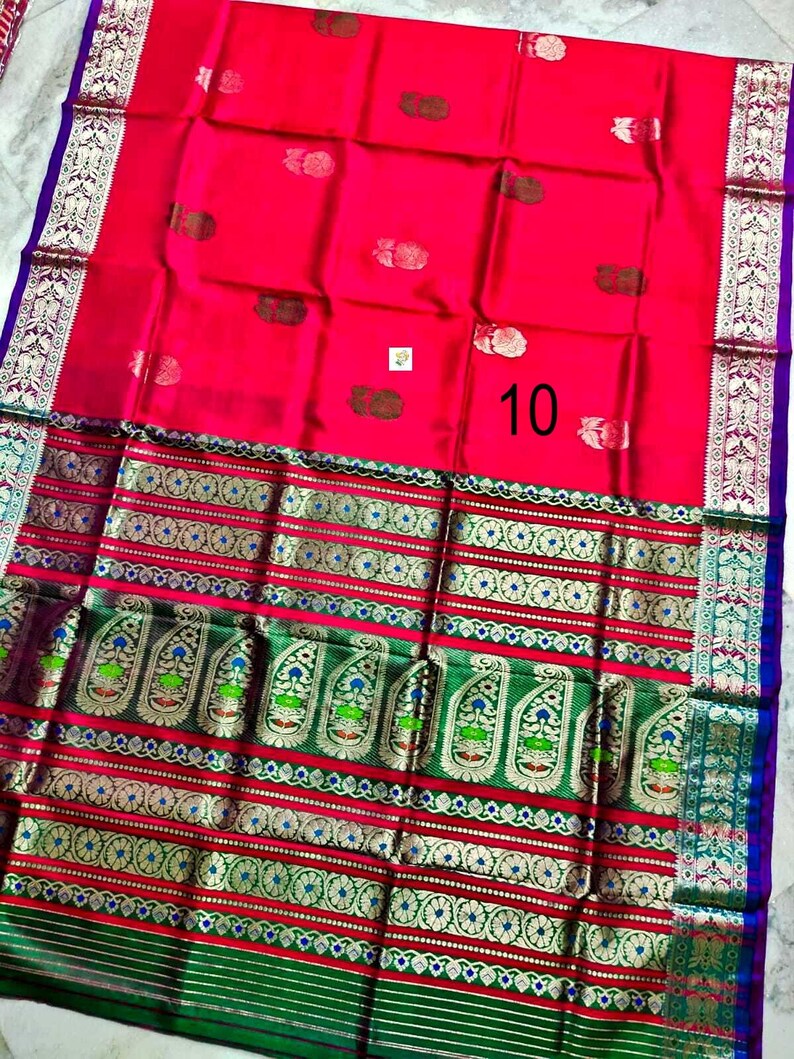 May include: A vibrant red saree featuring a wide green border with intricate gold and multicolored paisley patterns. The fabric displays small floral motifs across the main body and a prominent number 10 printed on the surface. The edges are finished with a purple trim.