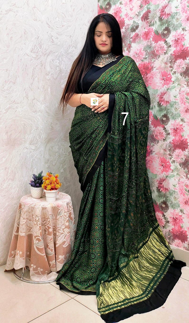 May include: A dark green saree featuring a repeating geometric pattern in black and gold, paired with a solid black blouse. The garment includes a wide, pleated gold metallic border at the hem and a silver-toned statement necklace.