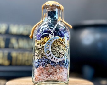 May include: A glass bottle filled with layers of amethyst crystals, dried herbs, lavender, and pink Himalayan salt. A silver crescent moon charm hangs from the top of the bottle, which is sealed with a gold wax.
