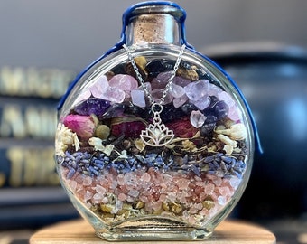 May include: A glass bottle filled with layers of dried flowers, herbs, and crystals. The bottle is tied with a blue ribbon and has a silver charm hanging from it. The bottle is sitting on a wooden base.